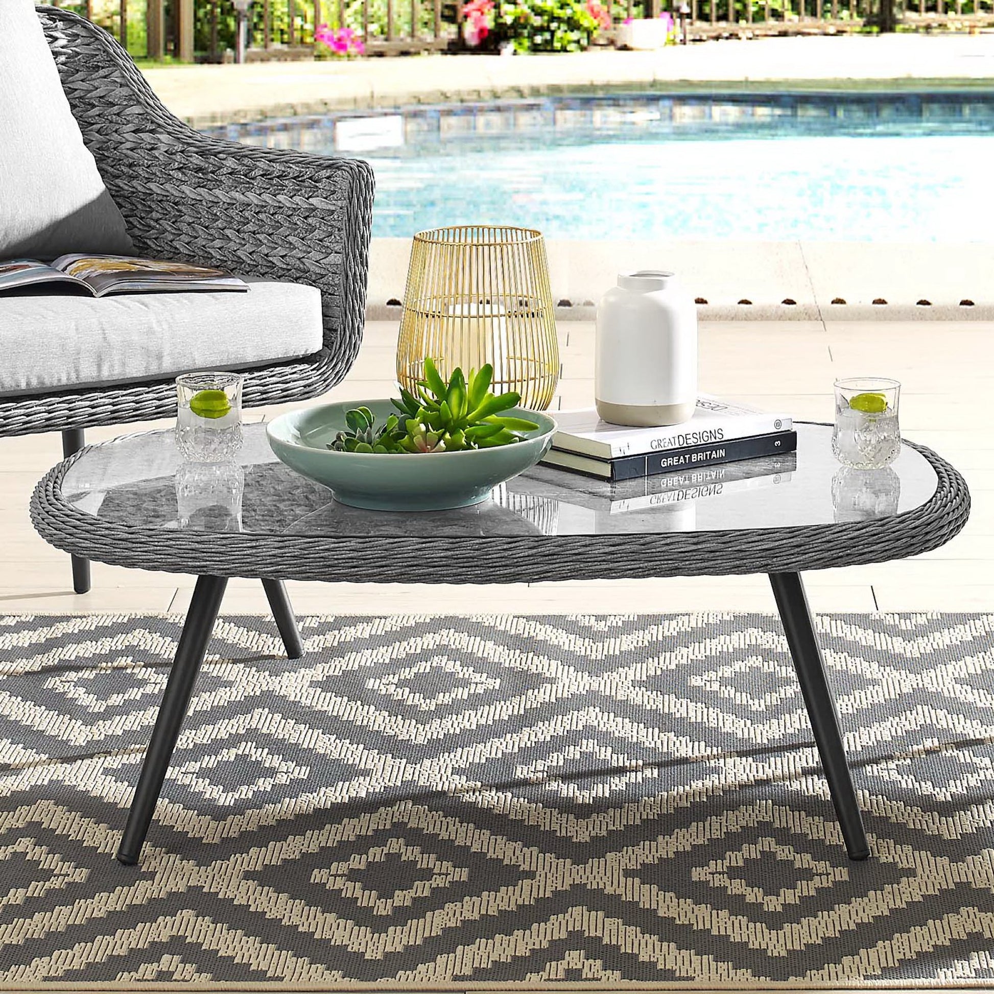 Gray woven rattan oval coffee table with glass top and black legs; succulent bowl, books, lantern, poolside patio.