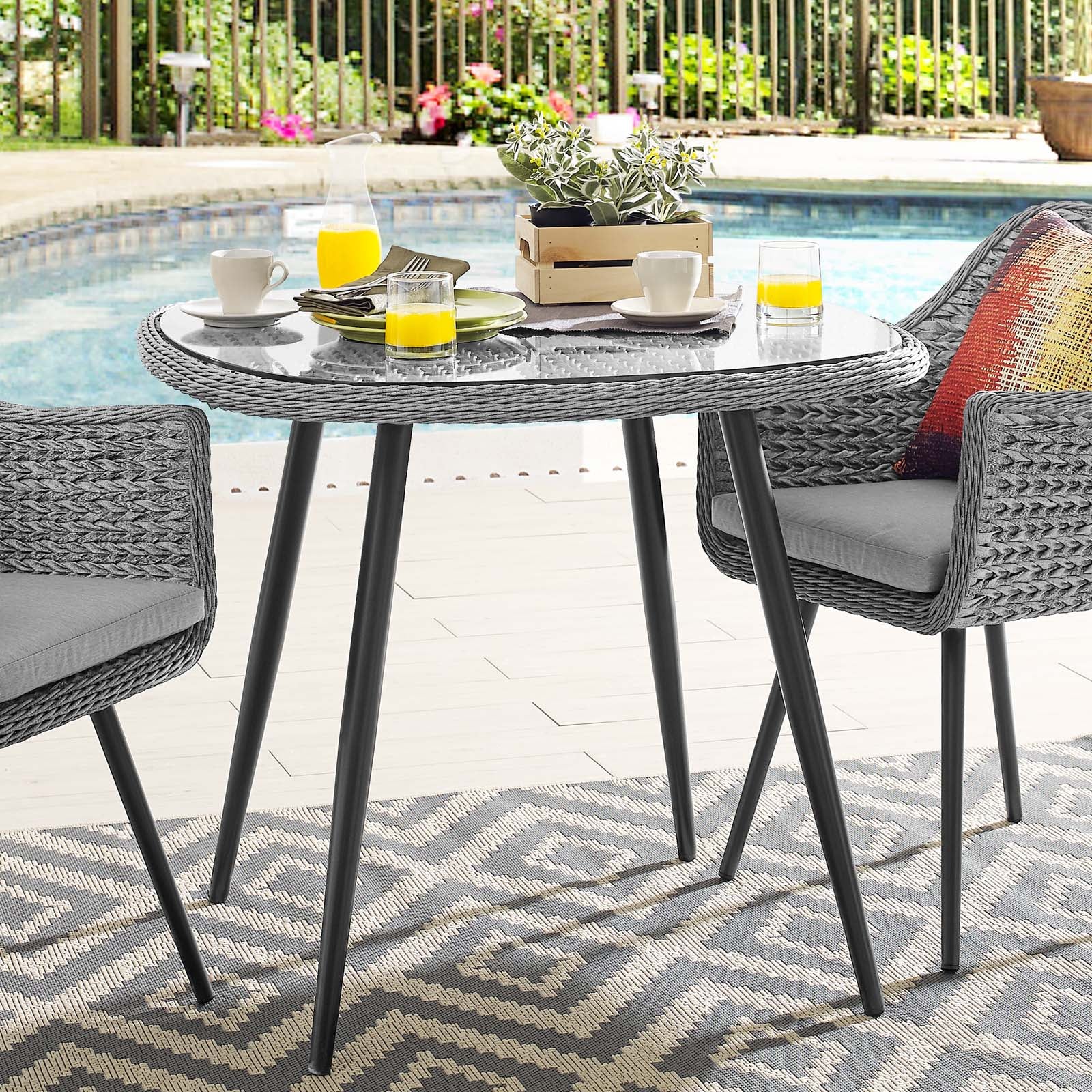 Grey round outdoor wicker table with glass top and black legs, two chairs; poolside breakfast with juice, cups, plant centerpiece.