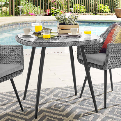 Grey round outdoor wicker table with glass top and black legs, two chairs; poolside breakfast with juice, cups, plant centerpiece.