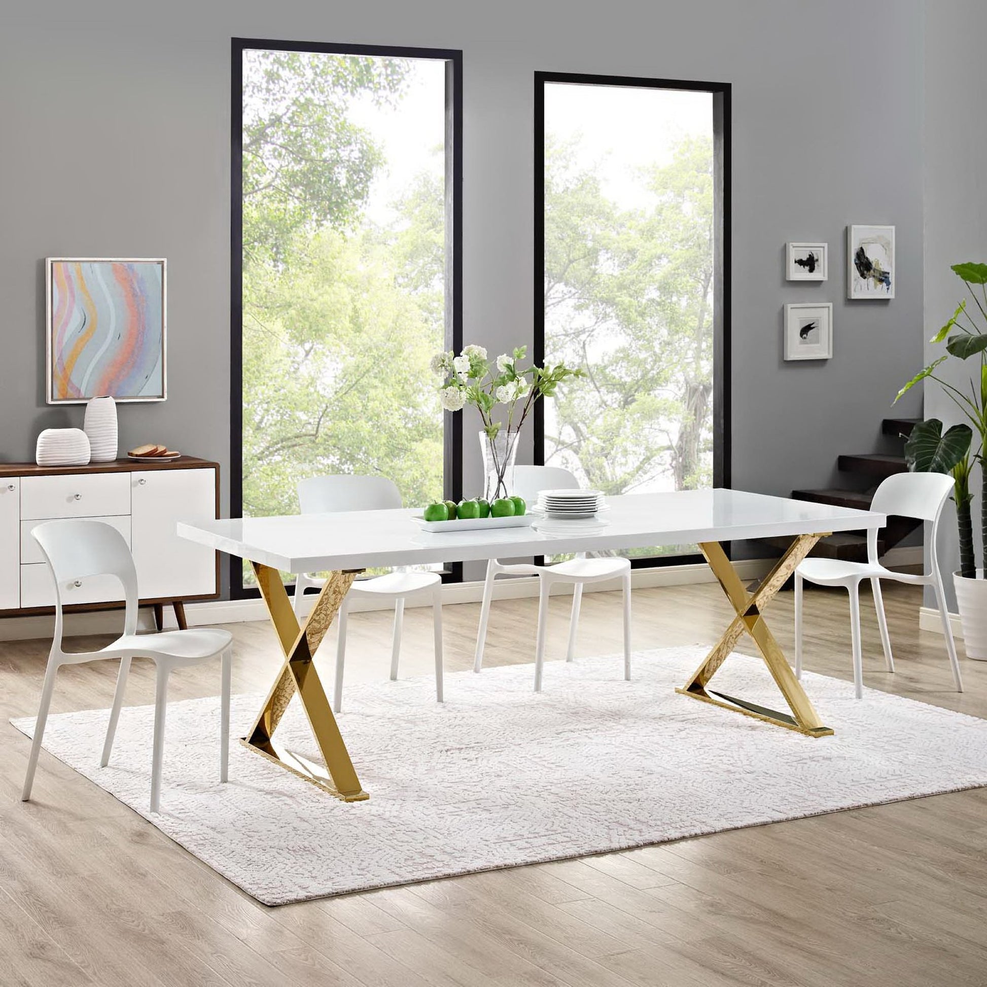 White rectangular dining table with gold X-frame legs by large windows.