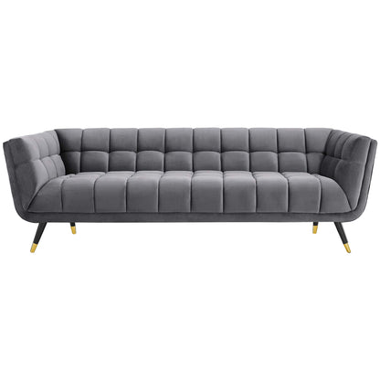 Gray velvet tufted sofa with grid cushions, low curved arms, angled legs with gold tips, modern mid-century style.