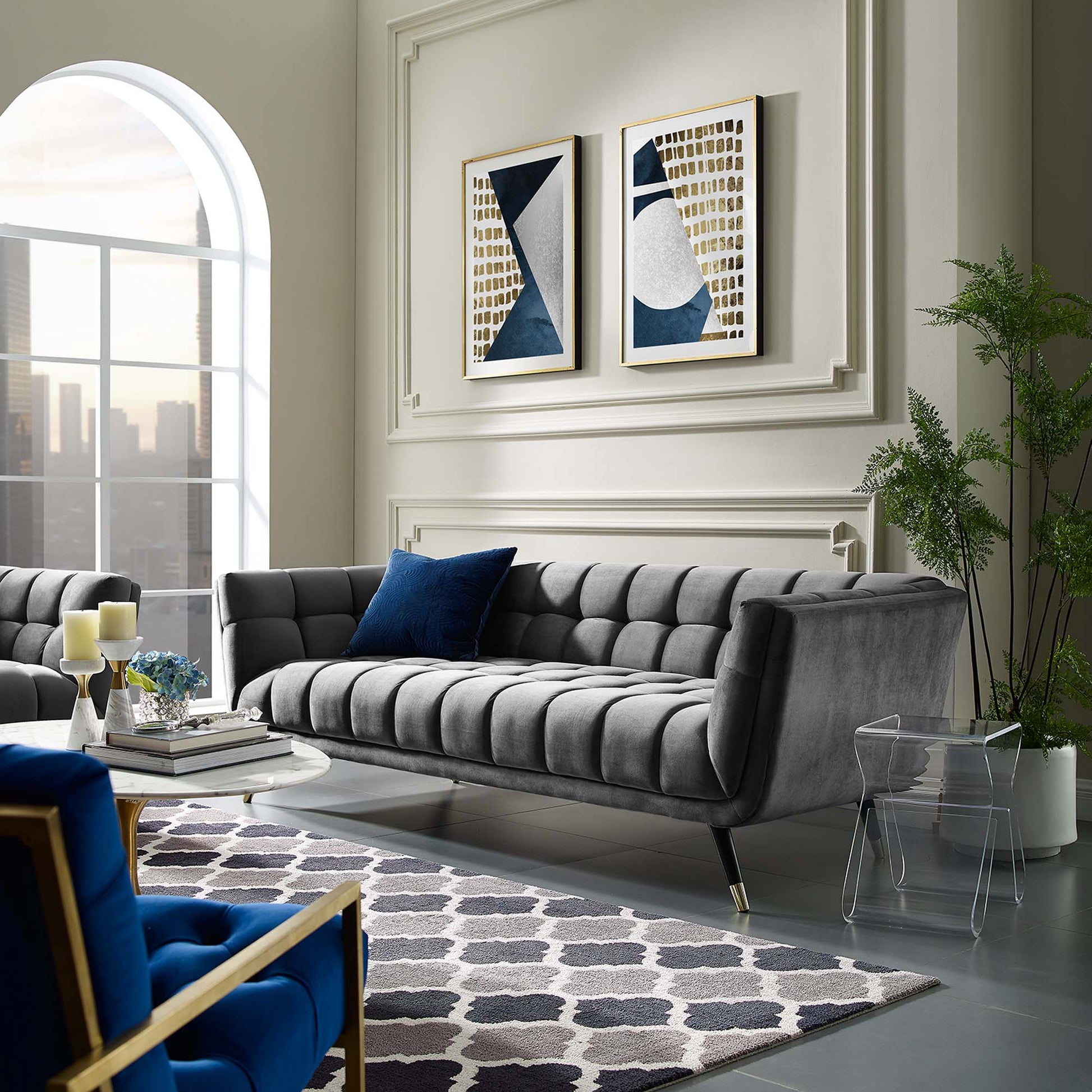 Gray tufted velvet sofa with blue pillow in a modern living room; gold-framed abstract art, arched window, glass chair, round coffee table, geometric rug.