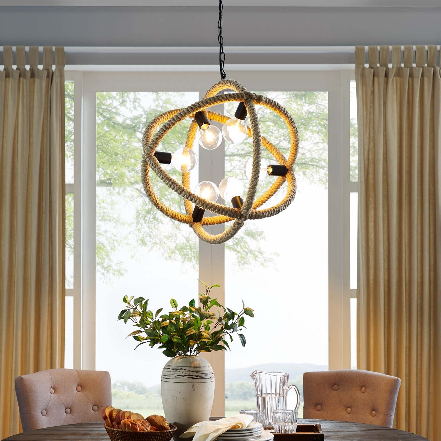 Natural rope pendant chandelier with exposed bulbs hangs over a wood dining table with tufted beige chairs and a greenery vase.