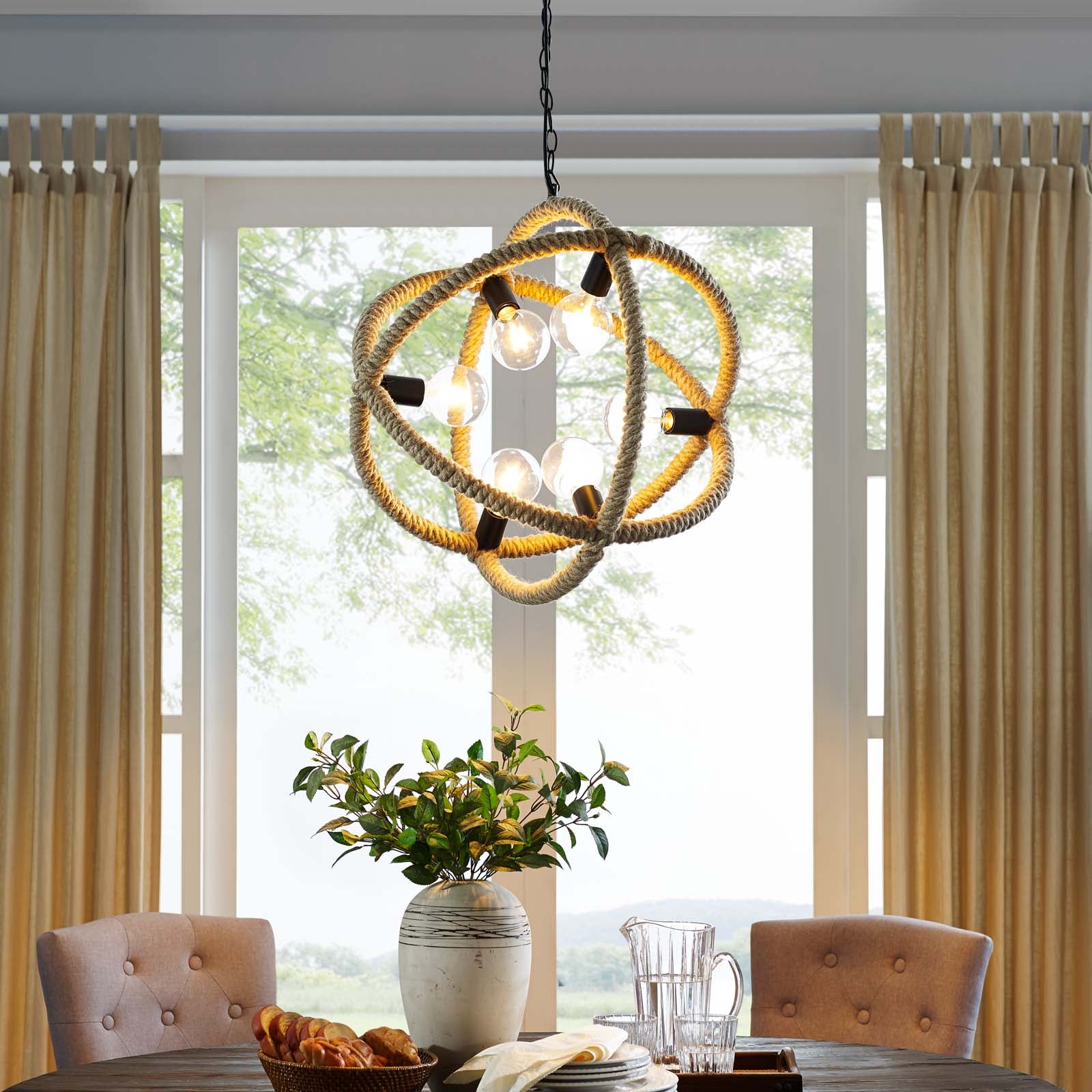 Natural rope pendant chandelier with exposed bulbs hangs over a wood dining table with tufted beige chairs and a greenery vase.