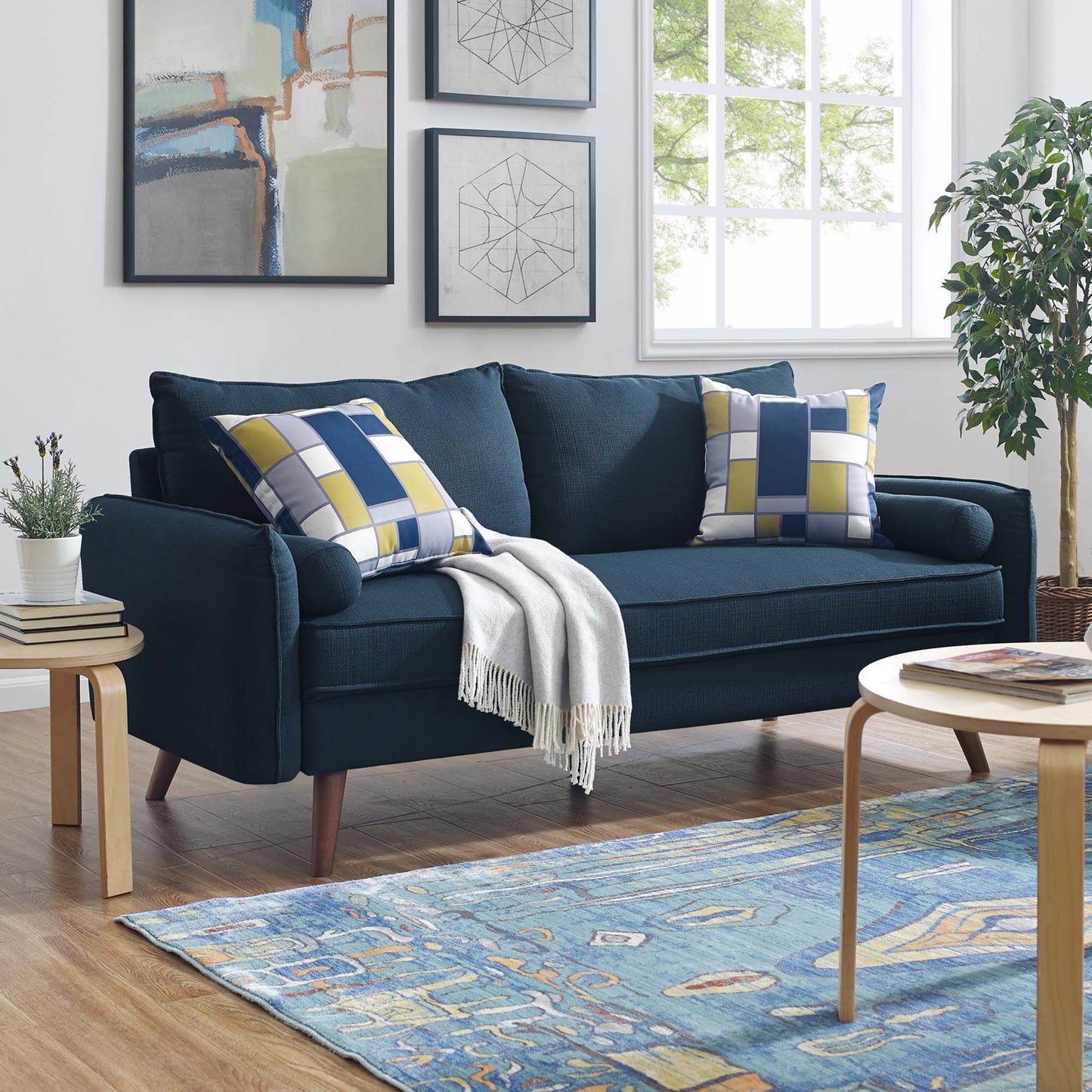 Dark blue sofa with yellow-blue plaid cushions, light throw, wooden coffee table, blue patterned rug, plant & wall art.