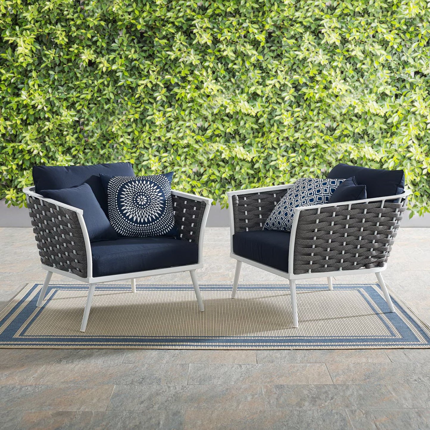 Pair of outdoor aluminum armchairs with white frames and navy cushions on a beige-blue striped rug beside a green hedge.