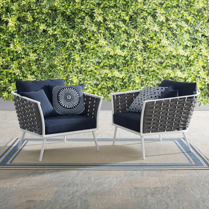 Pair of outdoor aluminum armchairs with white frames and navy cushions on a beige-blue striped rug beside a green hedge.