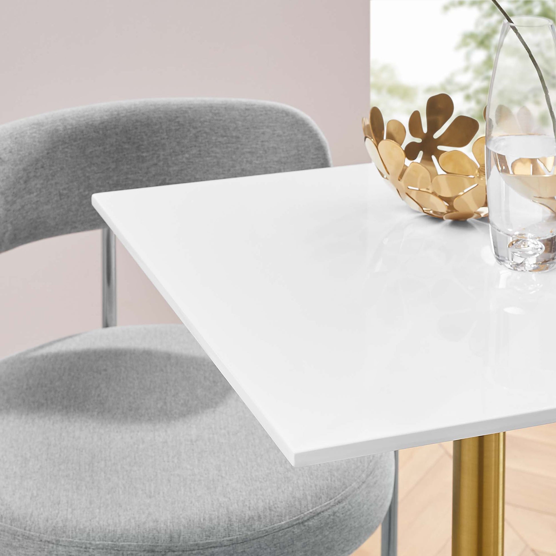 White square wood dining table with gold legs, paired with a grey upholstered chair & a gold decorative bowl.