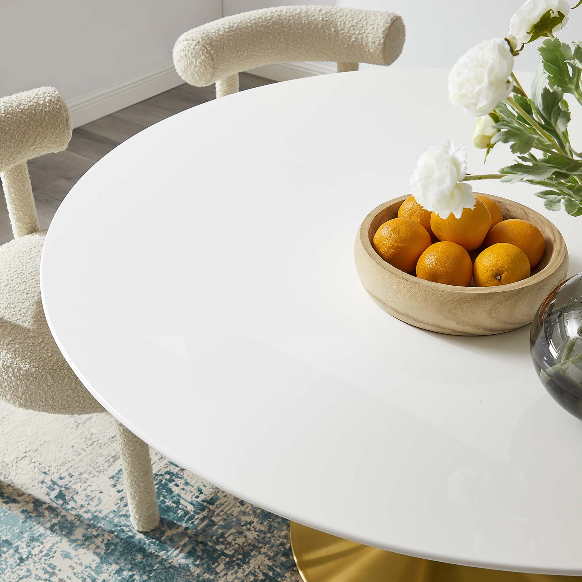 Round wooden dining table with a wooden bowl of oranges and white flowers in a glass vase; cream boucle chairs.
