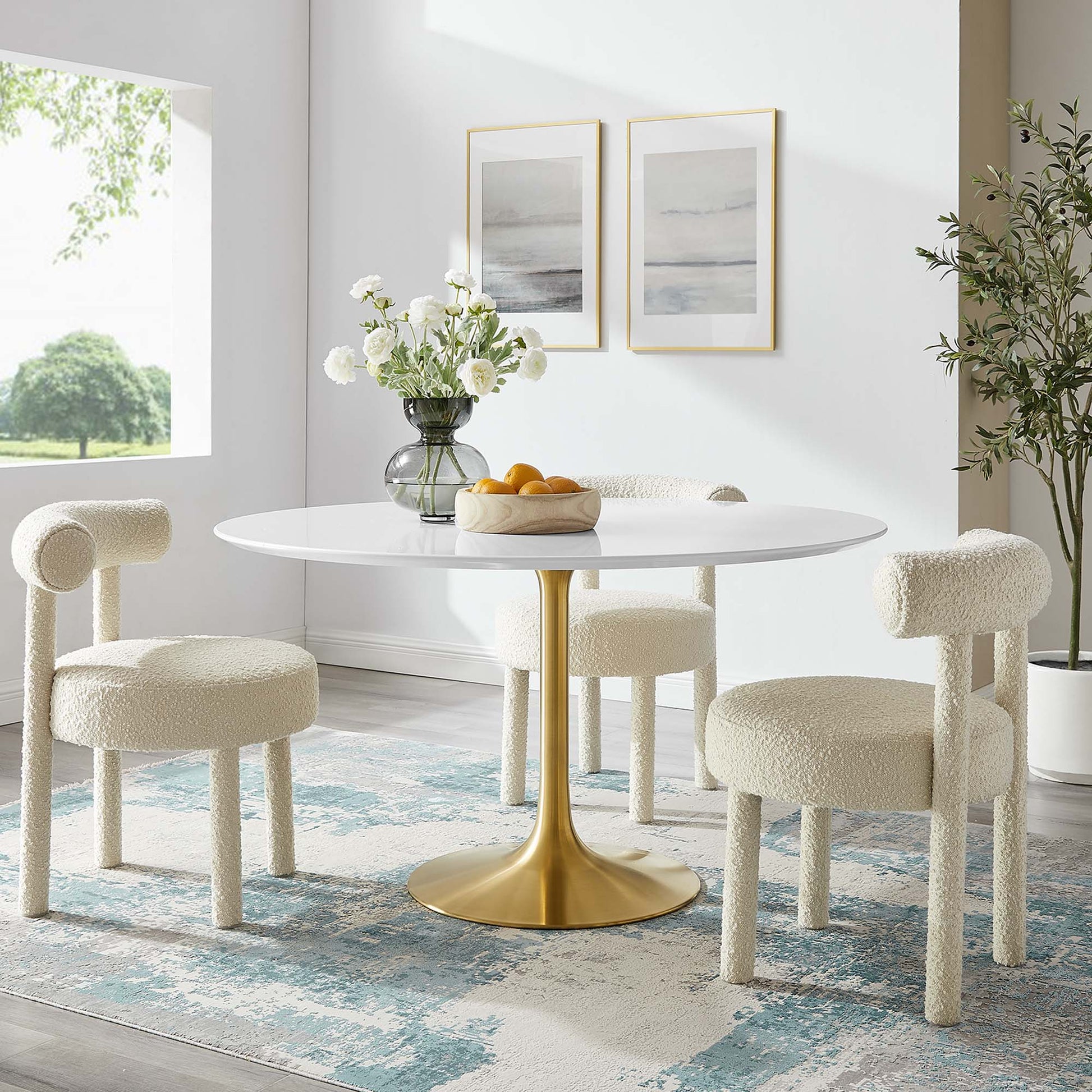 Round white dining table with gold pedestal, cream boucle chairs; vase of white flowers and oranges, blue rug.