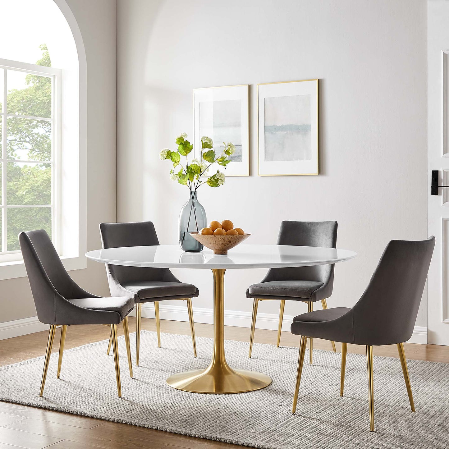 Round wood dining table with a gold pedestal, four gray chairs with gold legs, and a vase of green branches and oranges.