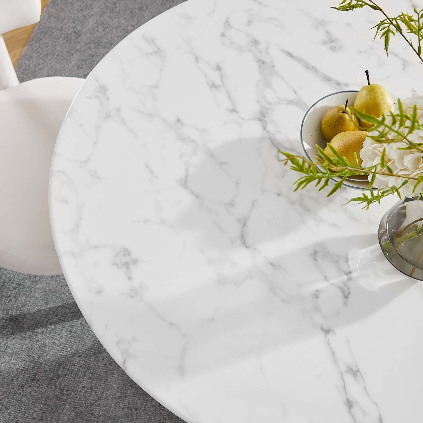 Round faux marble dining table with gray veining, topped with a bowl of pears and a glass vase with greenery, with a gray rug and white seating visible.