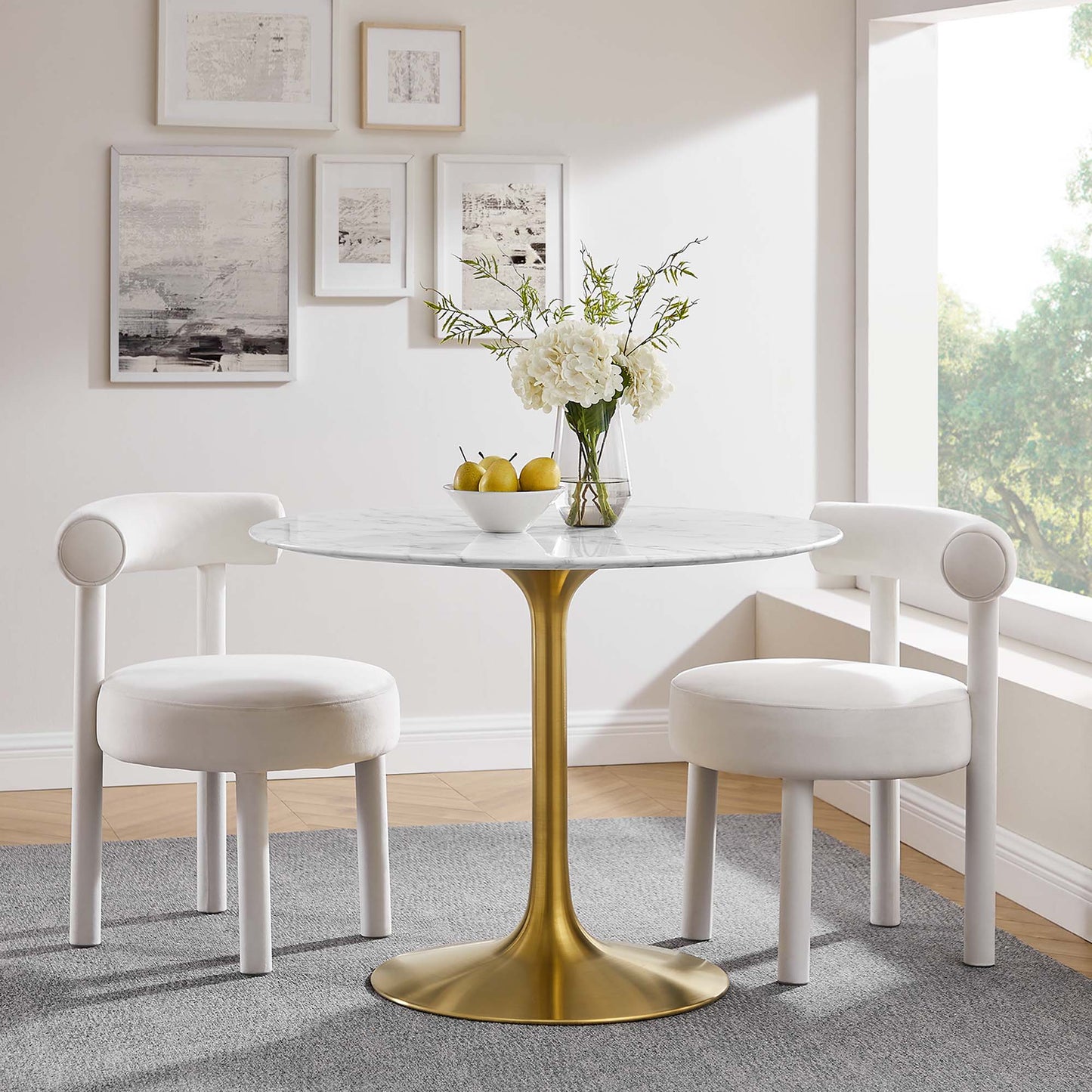 Round marble-top table with a gold pedestal, white upholstered chairs, vase of white flowers and lemons.