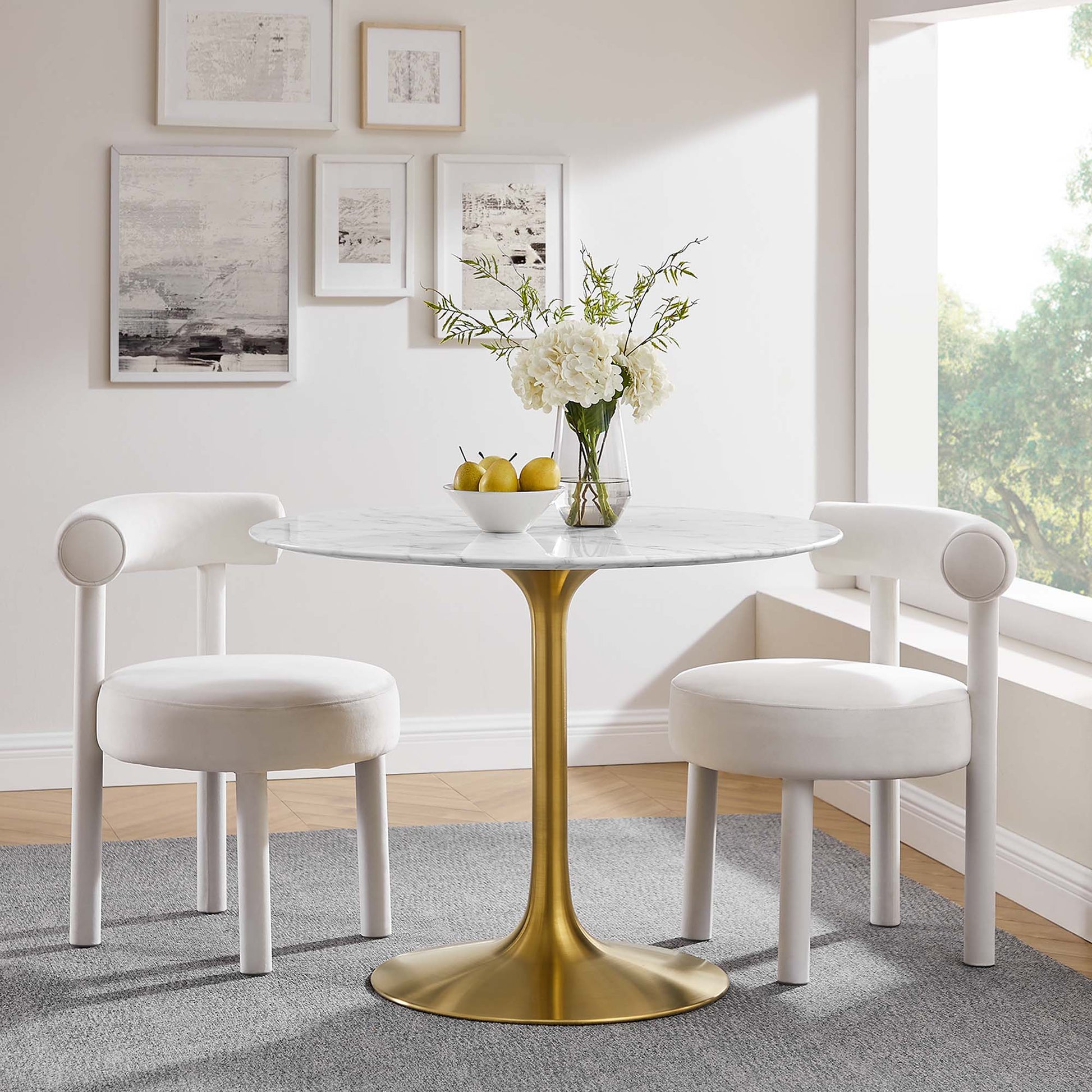 Round marble-top table with a gold pedestal, white upholstered chairs, vase of white flowers and lemons.