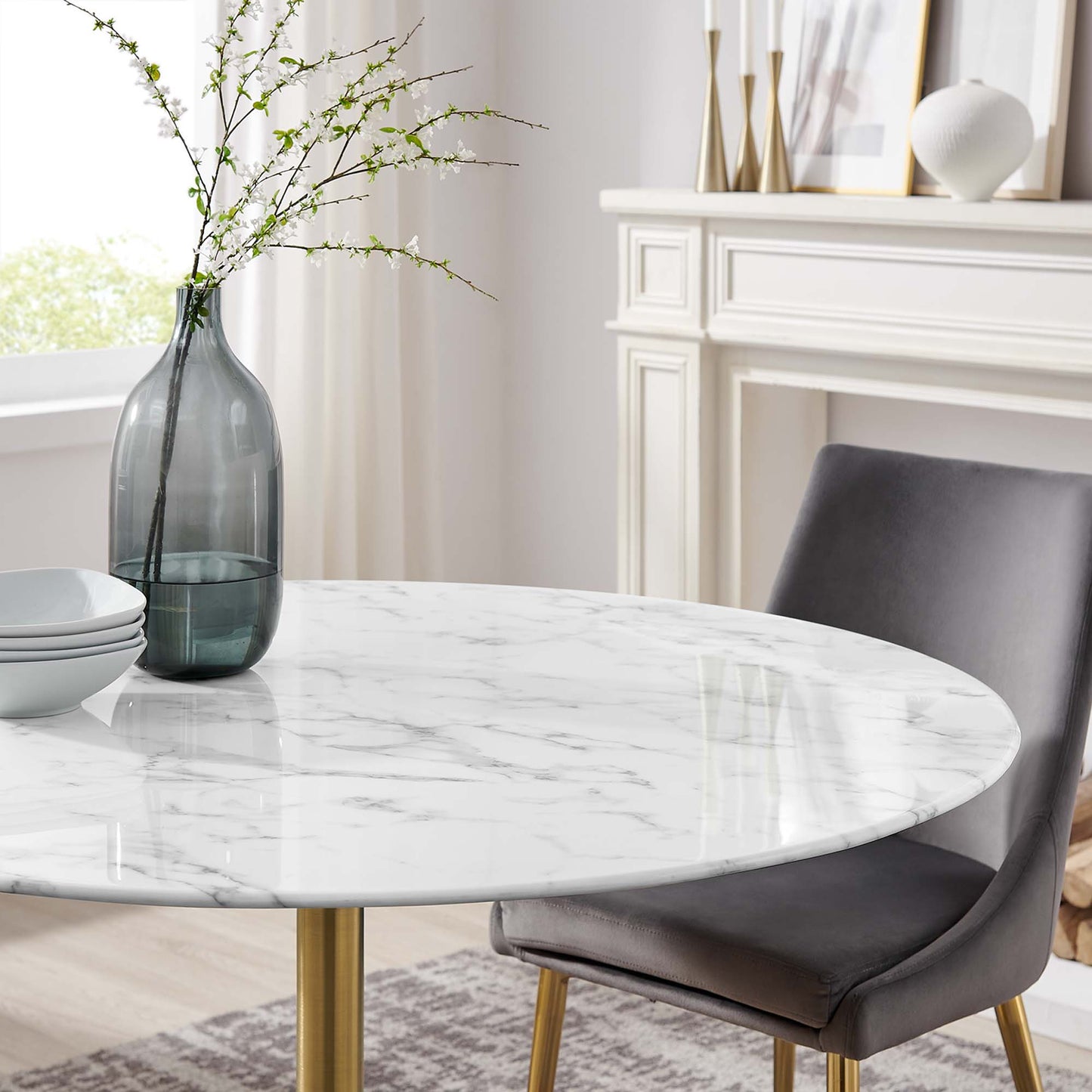 Round faux white marble dining table with gold metal legs in a bright room, gray chair nearby and smoky glass vase.