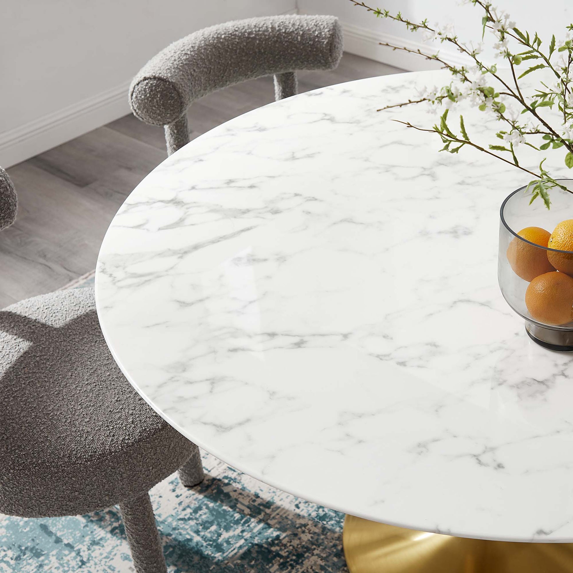 Round faux marble tabletop with a gold pedestal base, gray boucle chairs around it, and oranges in a glass bowl.