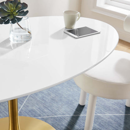 White oval wood dining table with a plant in a glass vase, a white mug and a tablet on top, on a blue rug with a white chair nearby.