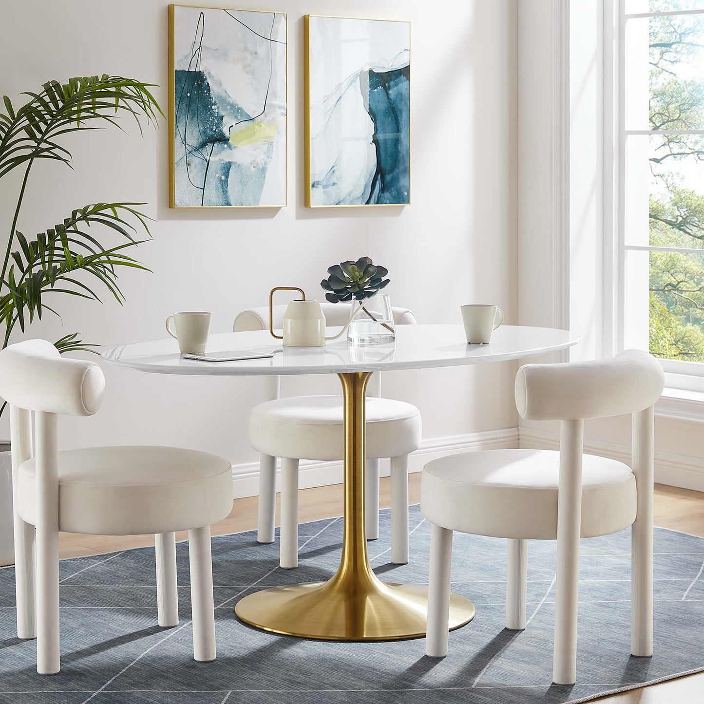 White oval wood dining table with a gold tulip pedestal and white upholstered chairs in a bright modern dining room with blue art.