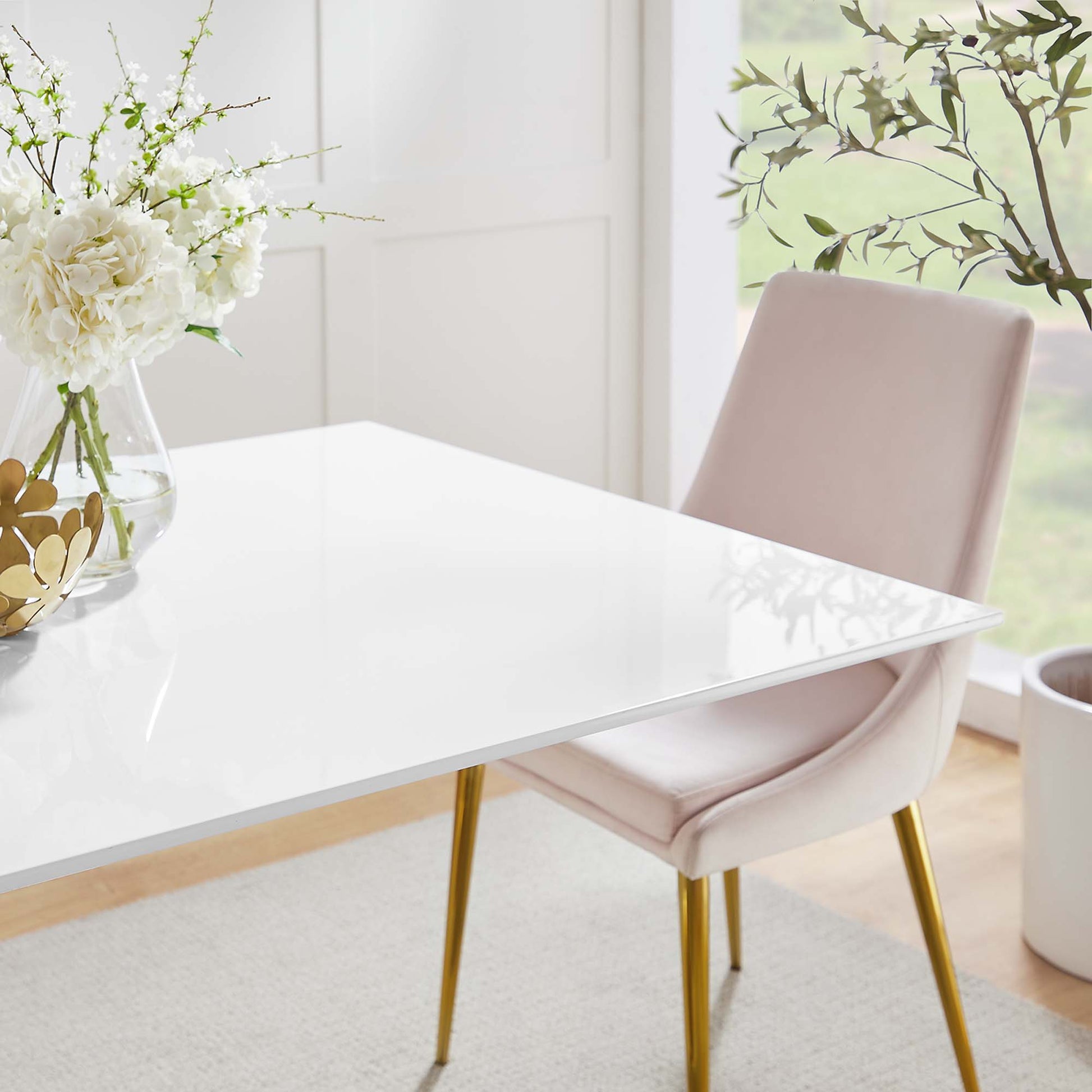 Glossy white table with gold legs, blush pink upholstered chair, vase of white flowers in bright airy dining area