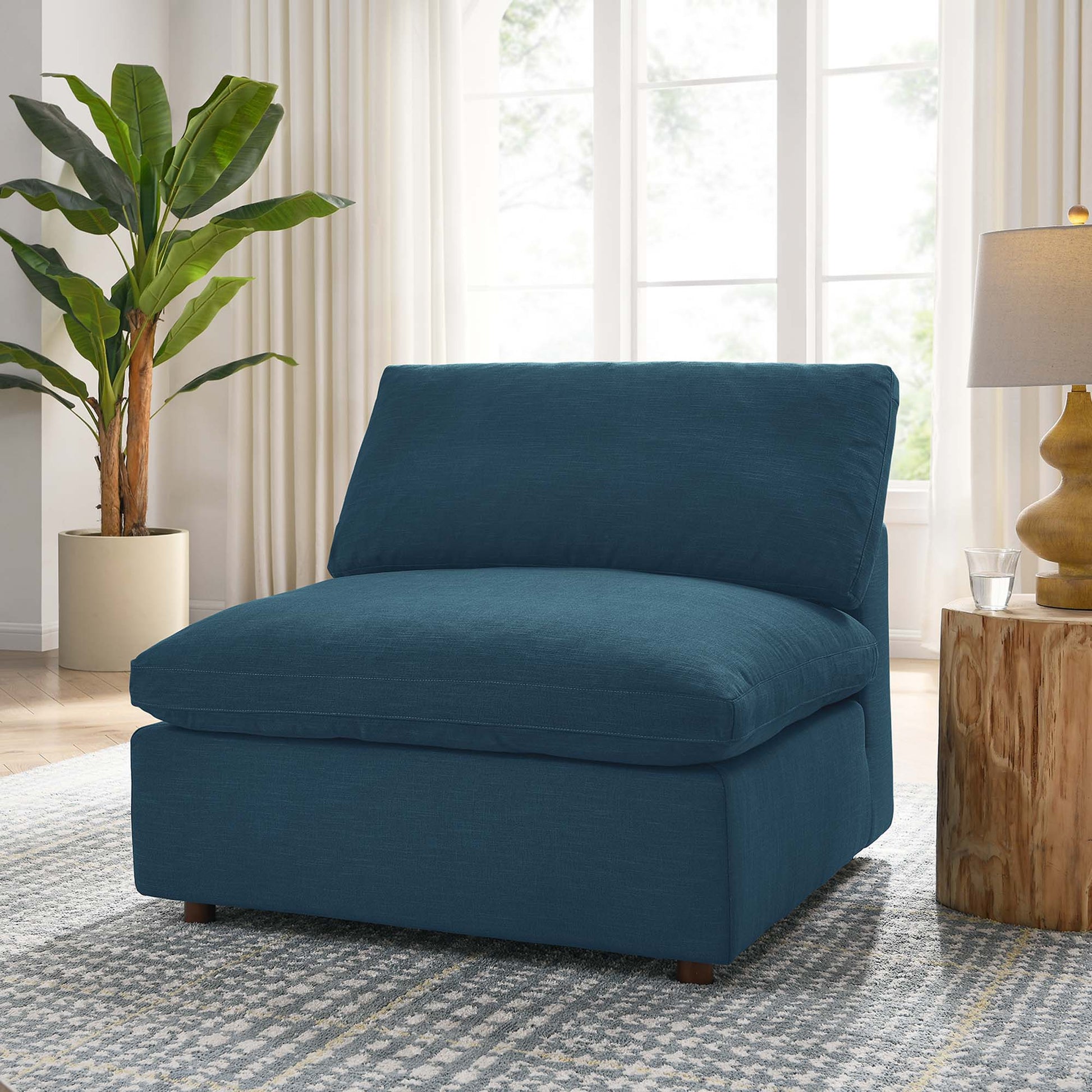 Blue armless upholstered chair with thick cushion in a modern living room setting with rug, plant, and wood stump side table.