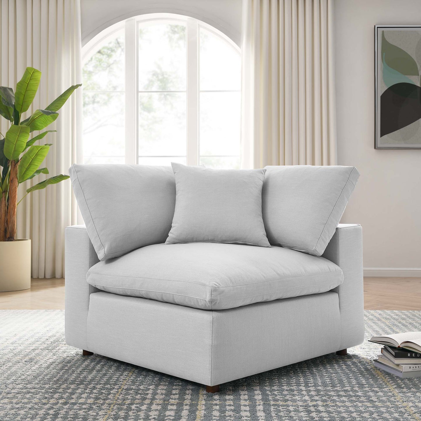 Light gray upholstered modern corner chair with a thick seat cushion and two back pillows in a living room with an arched window.