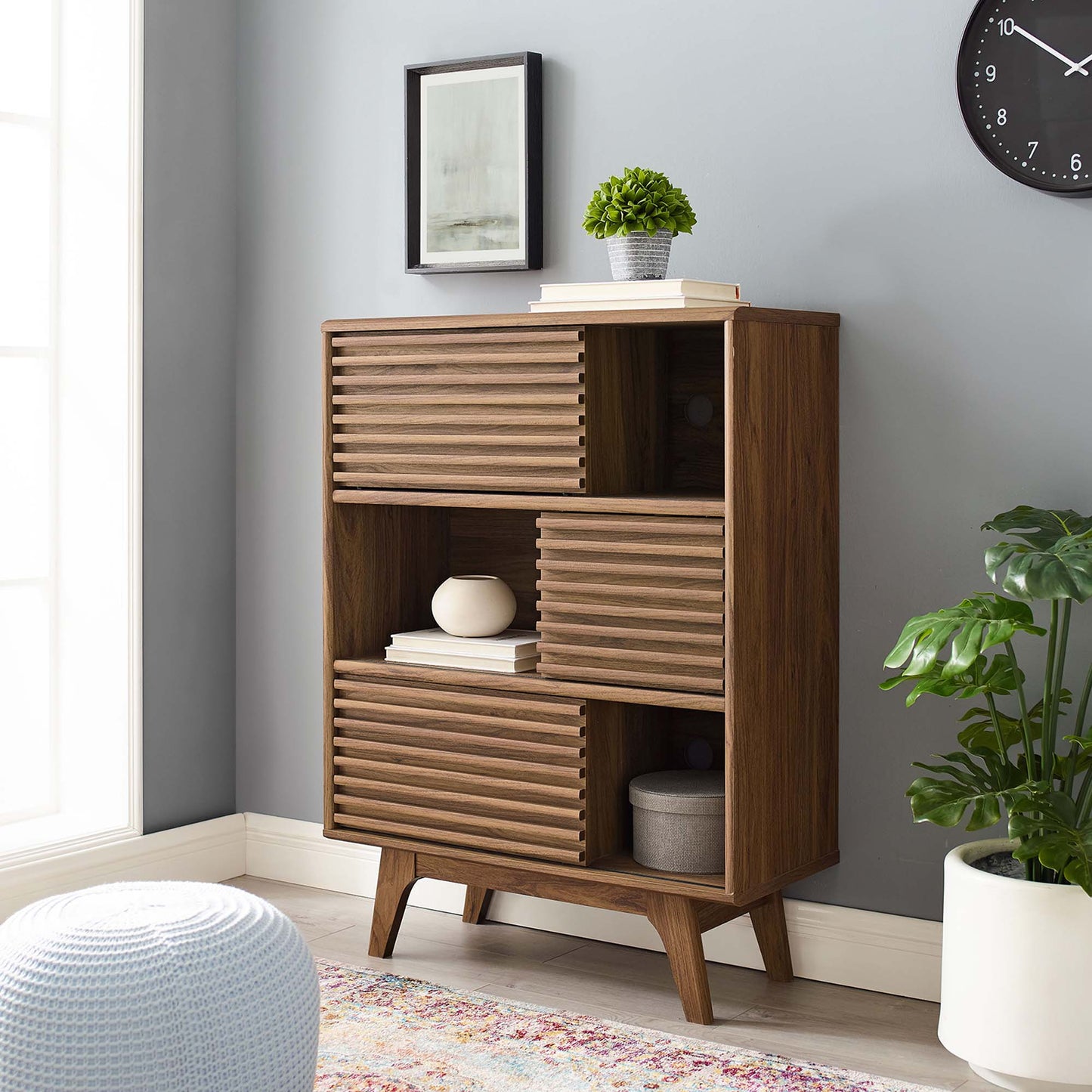 Walnut mid-century modern cabinet with slatted doors and open shelves, topped with a plant and books in a gray living room.