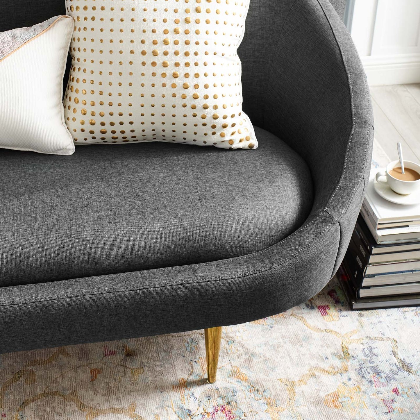Dark gray curved sofa with gold legs, white polka-dot pillow, alongside a stack of books and a coffee cup.