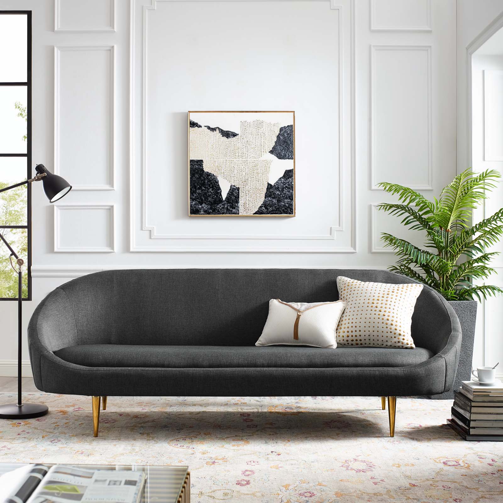 Charcoal gray curved sofa with gold legs & two decorative cushions in a bright modern living room with abstract wall art.