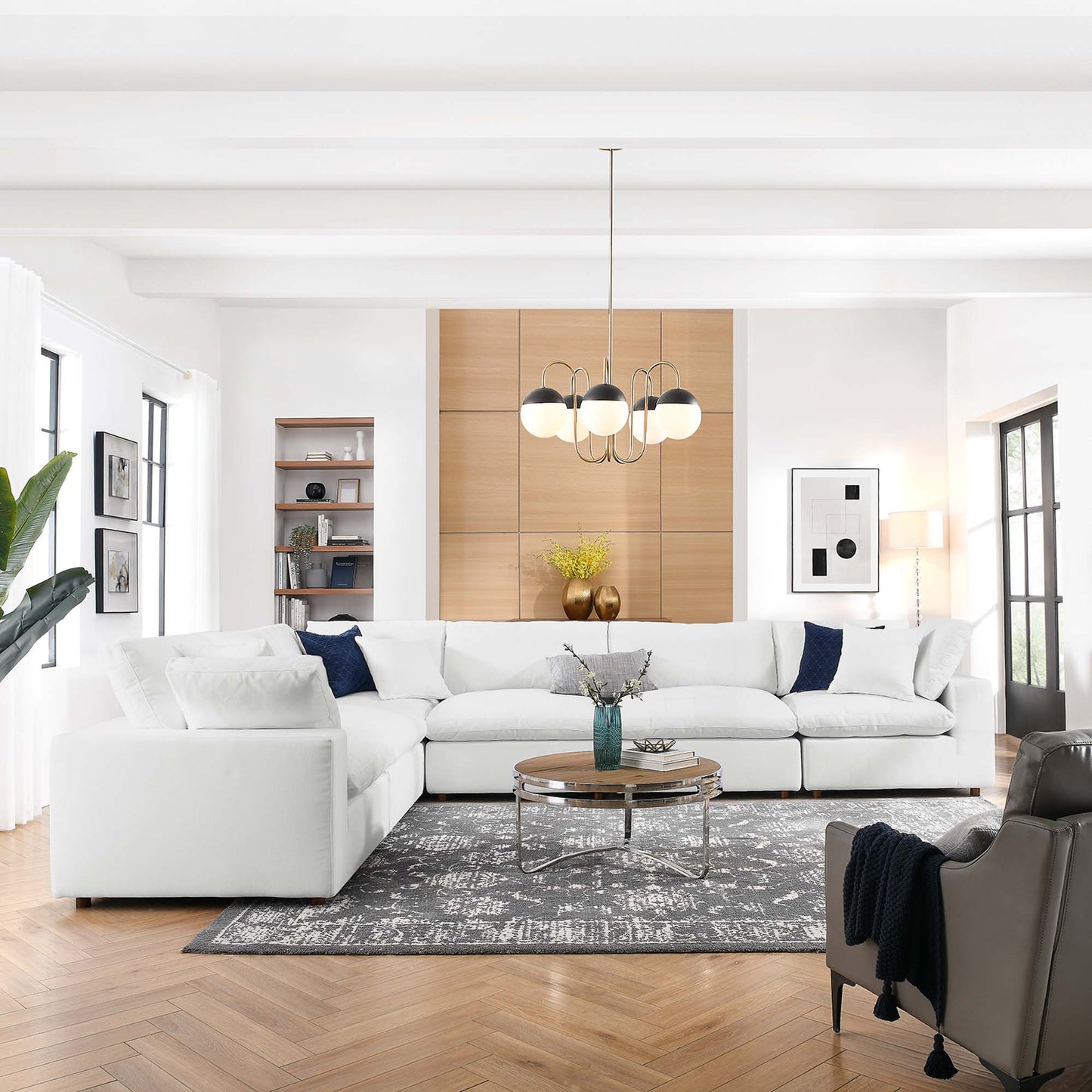 Bright modern living room with white sectional, navy pillows, round glass coffee table, gray rug, wood cabinet, chandelier.