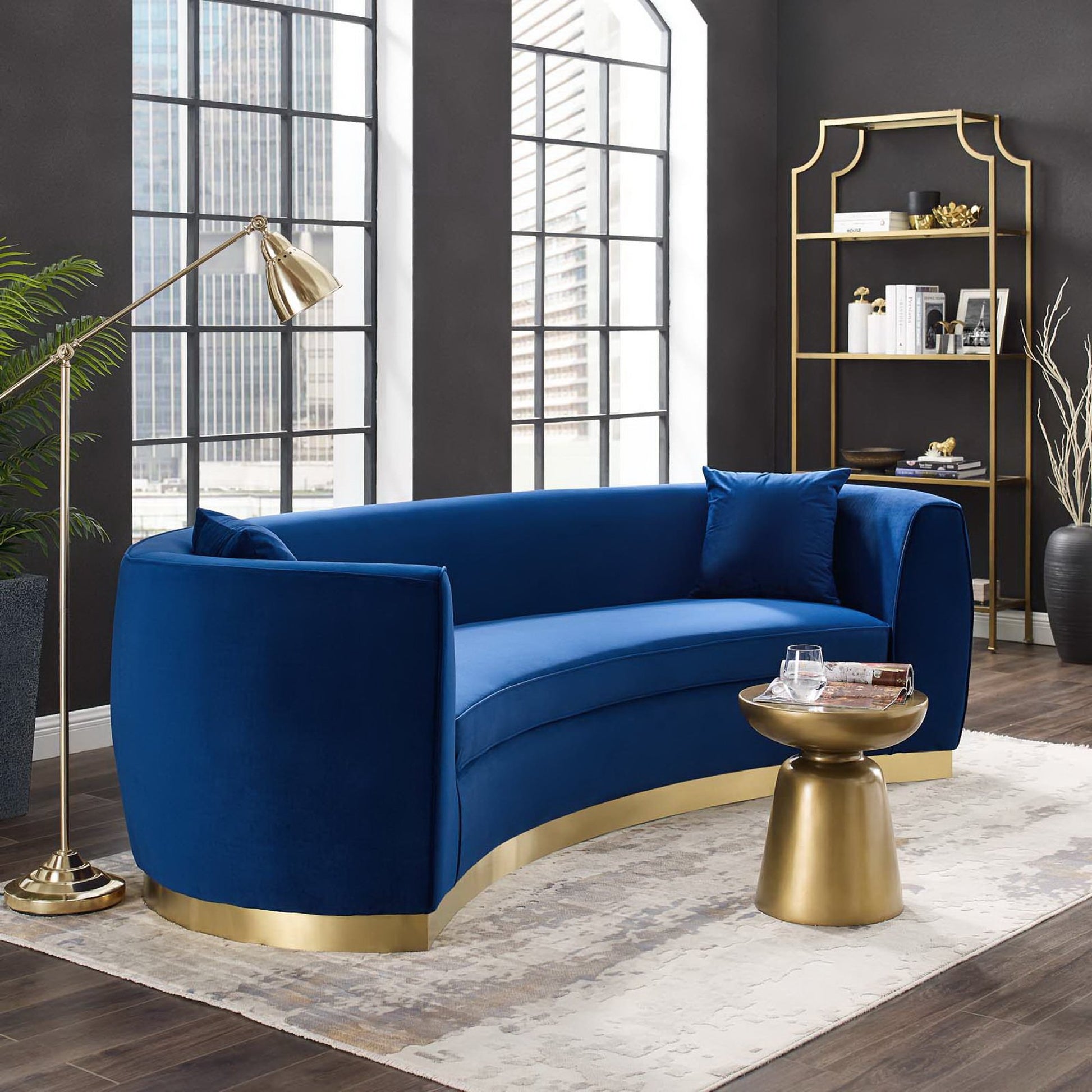 Navy velvet curved sofa with a gold base; gold floor lamp and round pedestal table; dark walls and large windows.