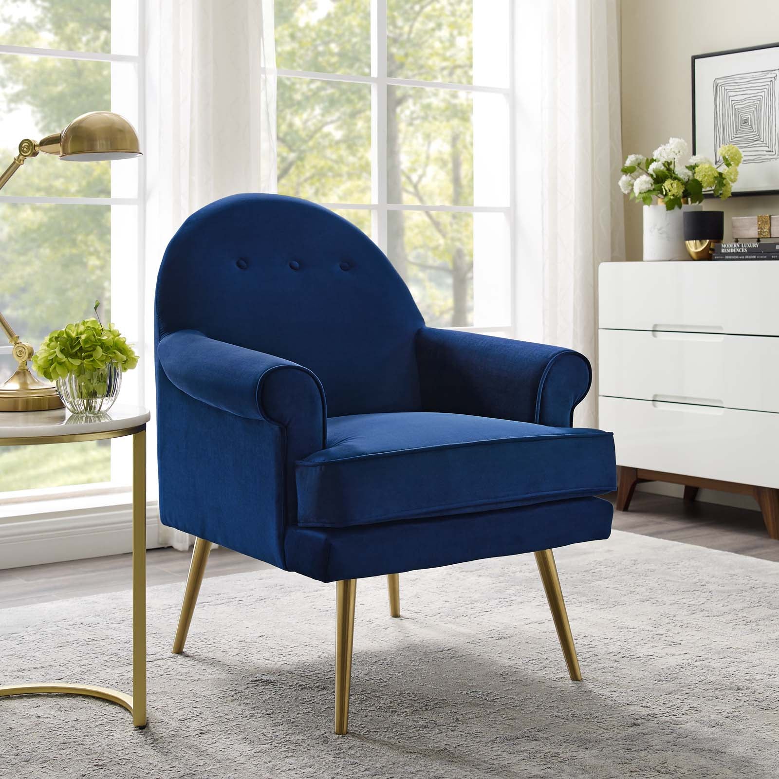 Blue velvet accent chair with gold legs, tufted back, and curved arms in a bright living room.