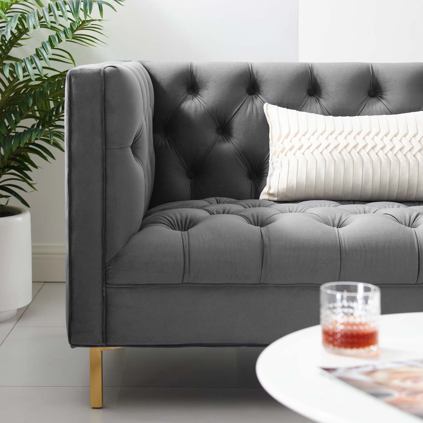 Grey tufted velvet sofa with button detailing, gold legs, cream striped pillow; plant & glass on white coffee table.