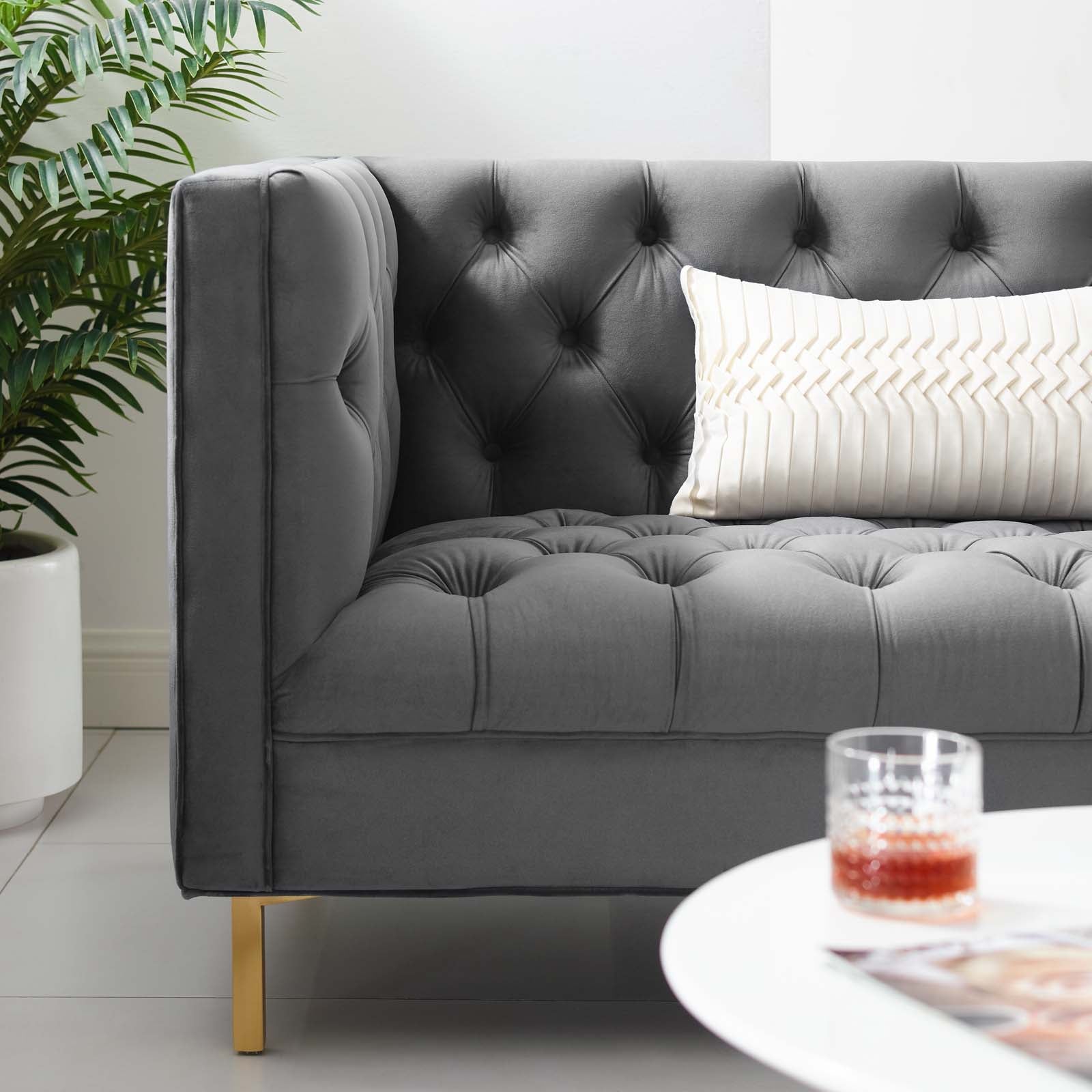 Grey tufted velvet sofa with button detailing, gold legs, cream striped pillow; plant & glass on white coffee table.