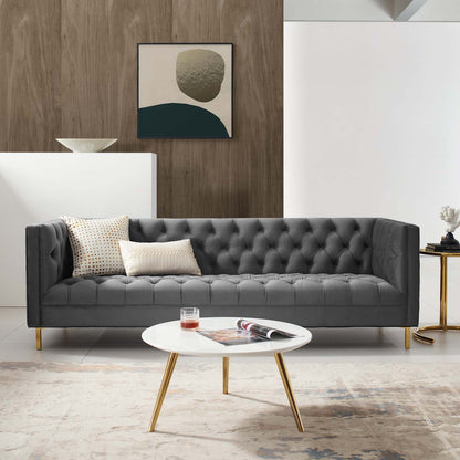 Dark gray tufted mid-century sofa with gold legs, cream pillows, white round coffee table, abstract art on wooden wall.