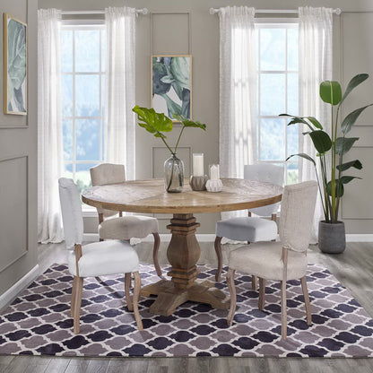 Round pine wood pedestal dining table with beige upholstered chairs; purple geometric trellis rug, leafy plants, and light gray walls.