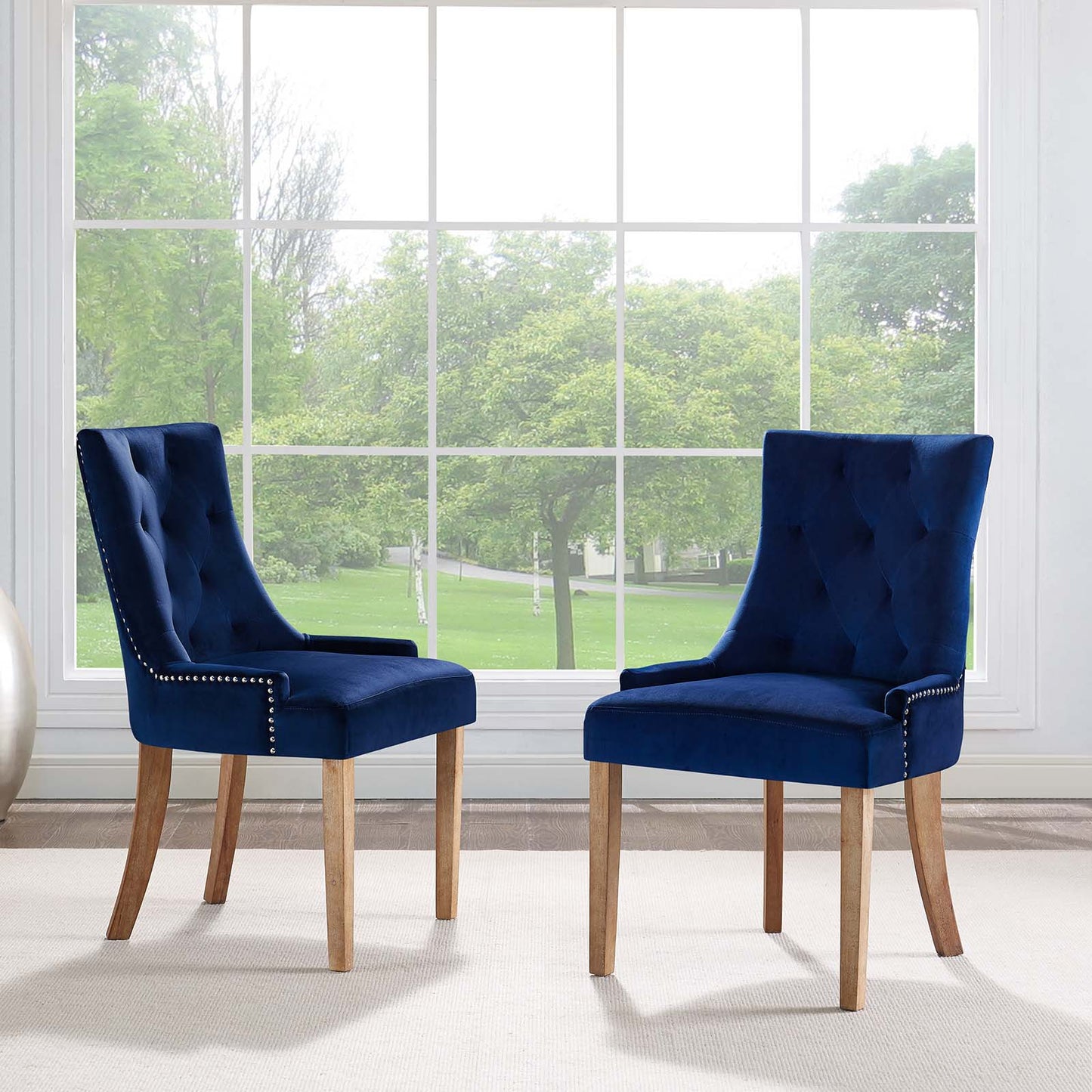 Two navy blue tufted velvet dining chairs with light wood legs and nailhead trim, in front of a large window.