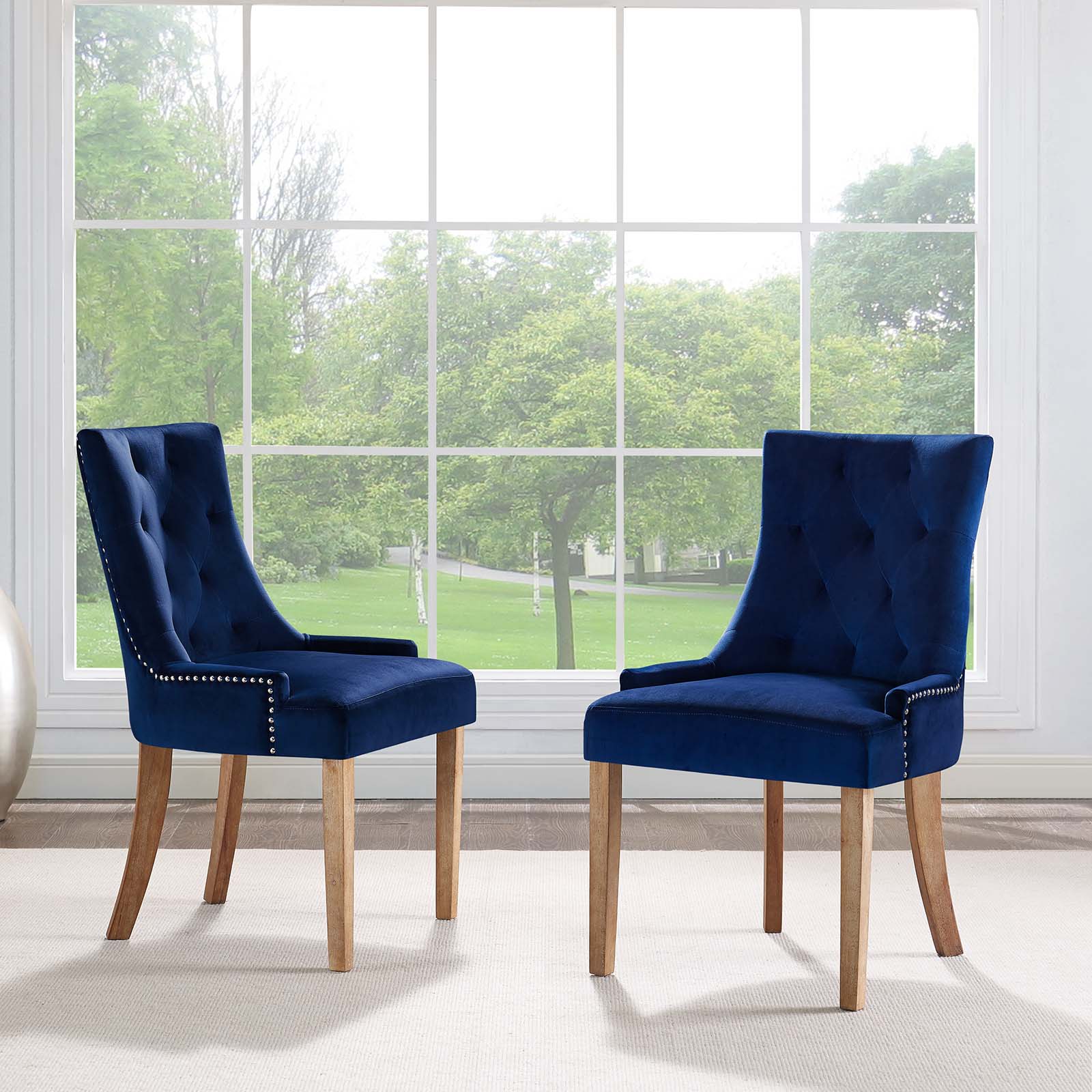Two navy blue tufted velvet dining chairs with light wood legs and nailhead trim, in front of a large window.