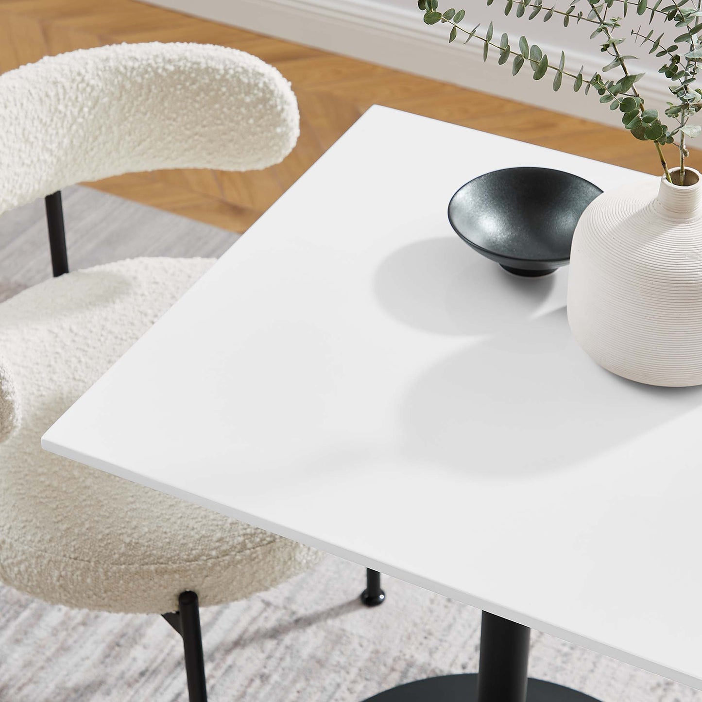 White square dining table with a small black bowl and a white vase with greenery; cream boucle chair nearby.