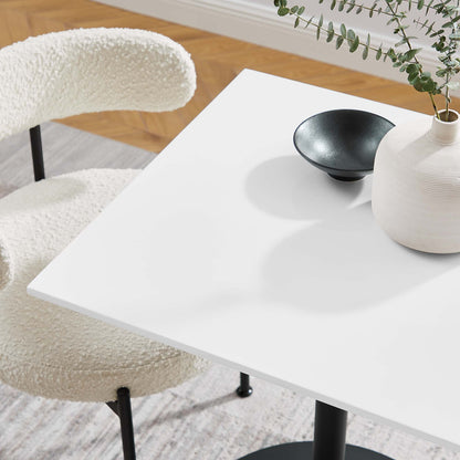 White square dining table with a small black bowl and a white vase with greenery; cream boucle chair nearby.