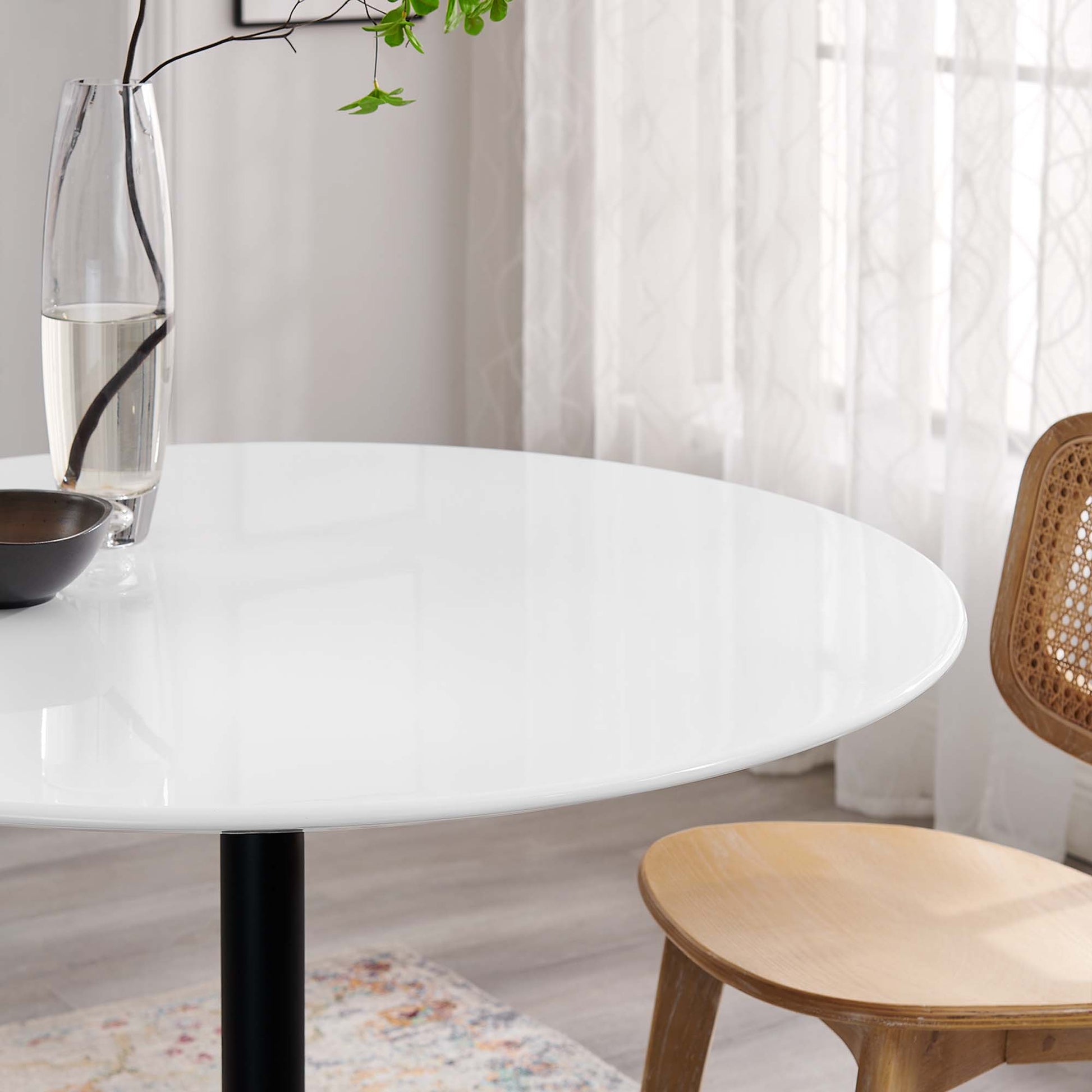 Round wood dining table with black pedestal leg; modern minimalist style, glass carafe, brown bowl, light-filled room.