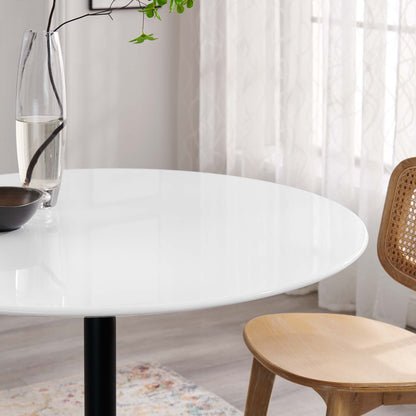 Round wood dining table with black pedestal leg; modern minimalist style, glass carafe, brown bowl, light-filled room.
