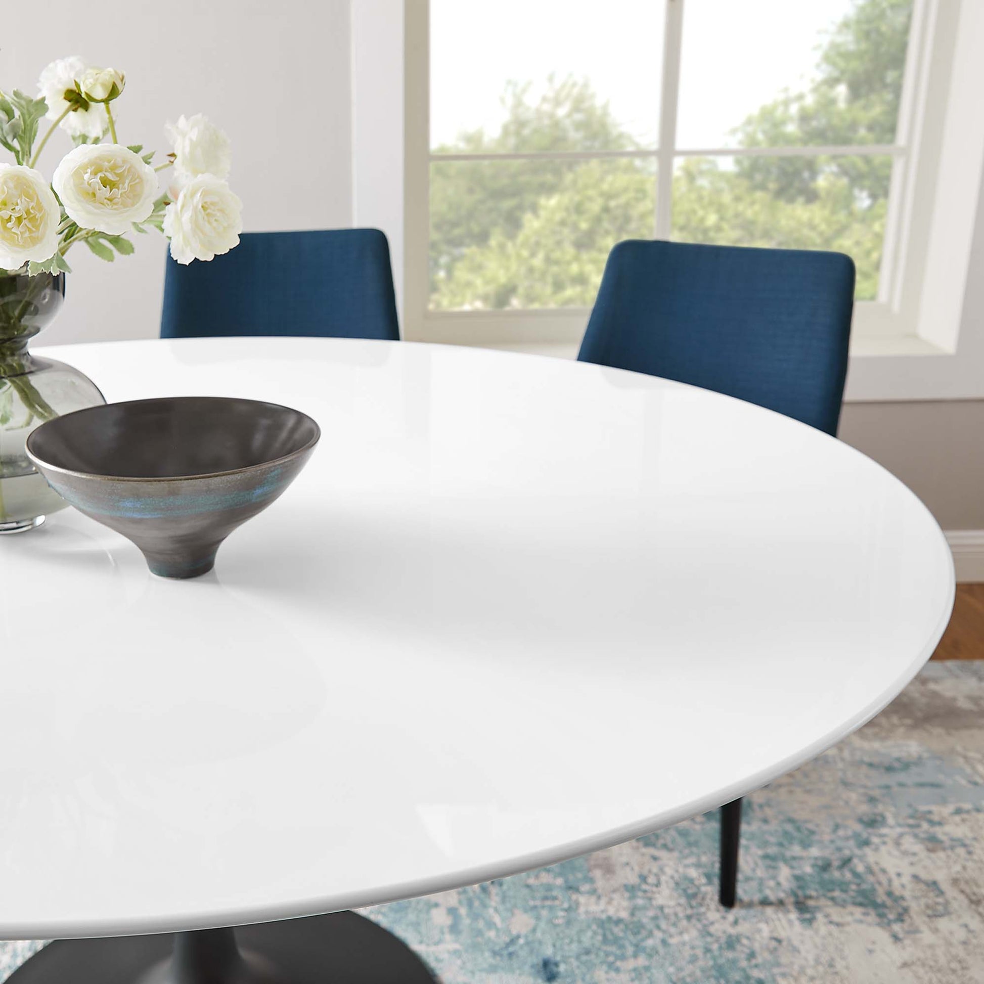Round wood dining table in white finish with a dark ceramic bowl, vase of white flowers, blue upholstered chairs, and a bright window view.