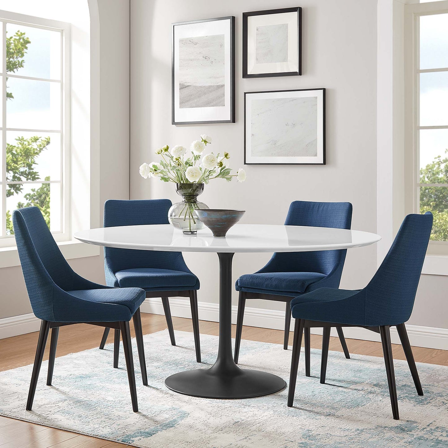 Round white pedestal dining table with four navy blue upholstered chairs, black base, vase of flowers, wall art.