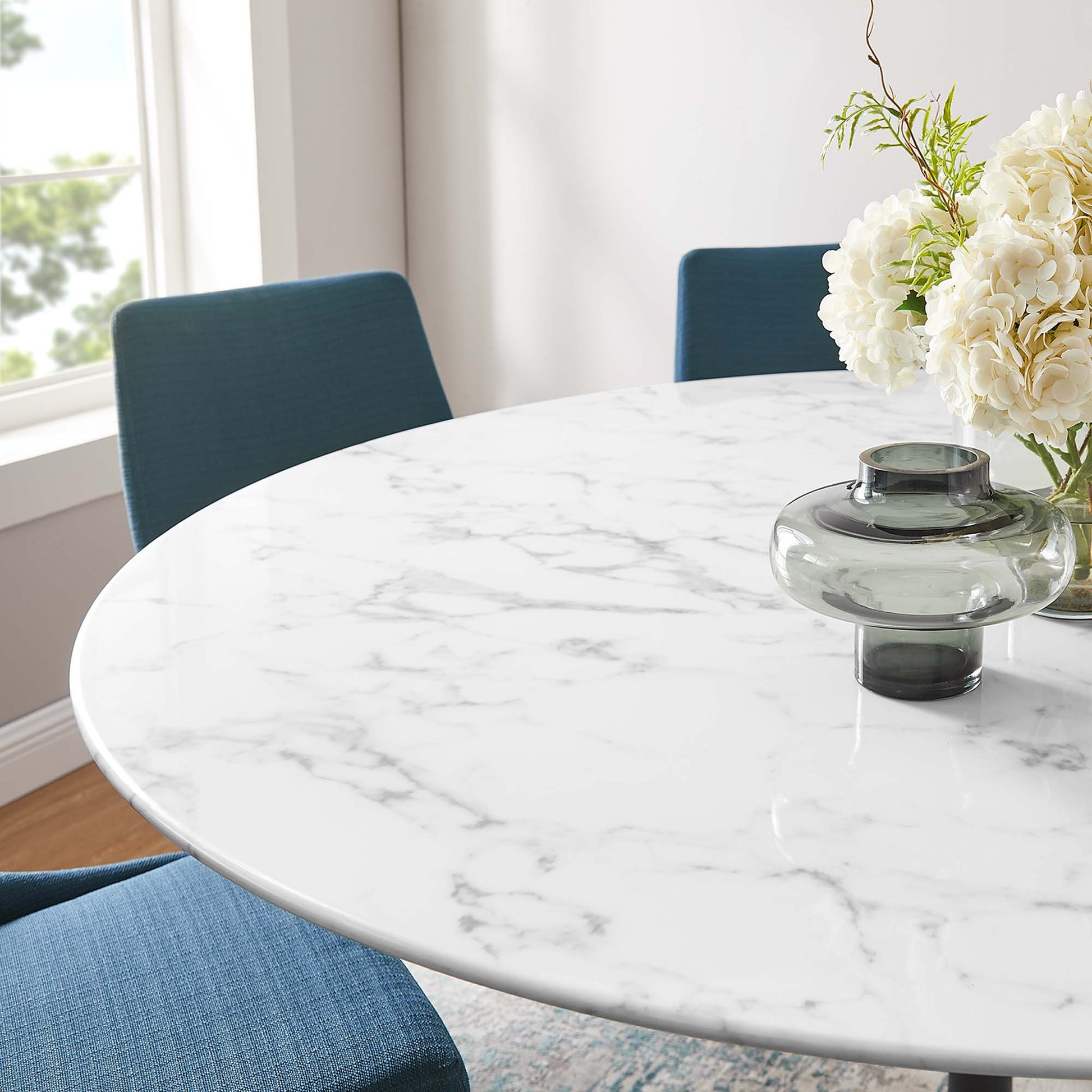 Round faux marble-top dining table with blue upholstered chairs and a glass vase centerpiece in a sunlit dining nook.