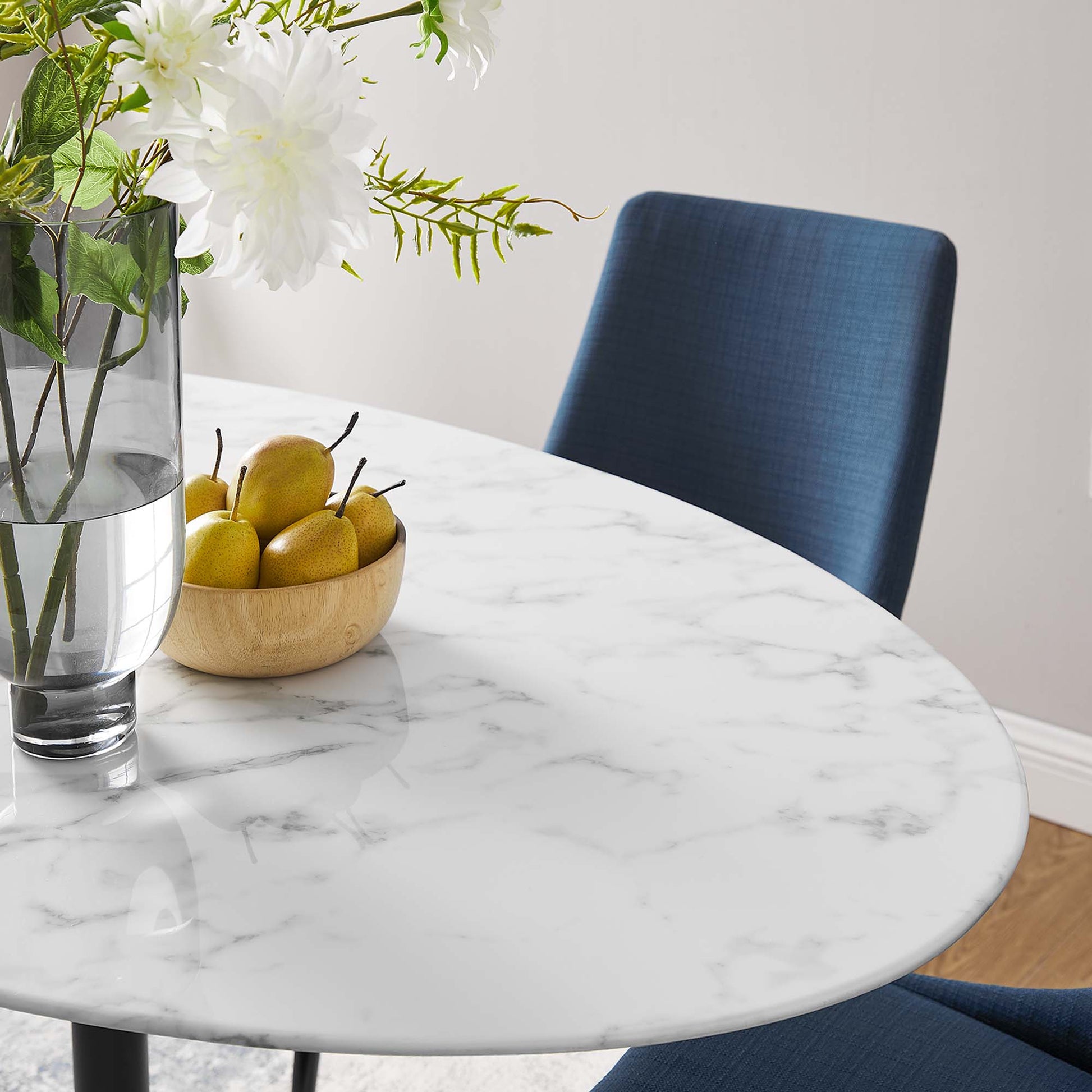 Oval faux marble dining table with gray veining, blue upholstered chair, vase of white flowers, and a wooden bowl of pears.
