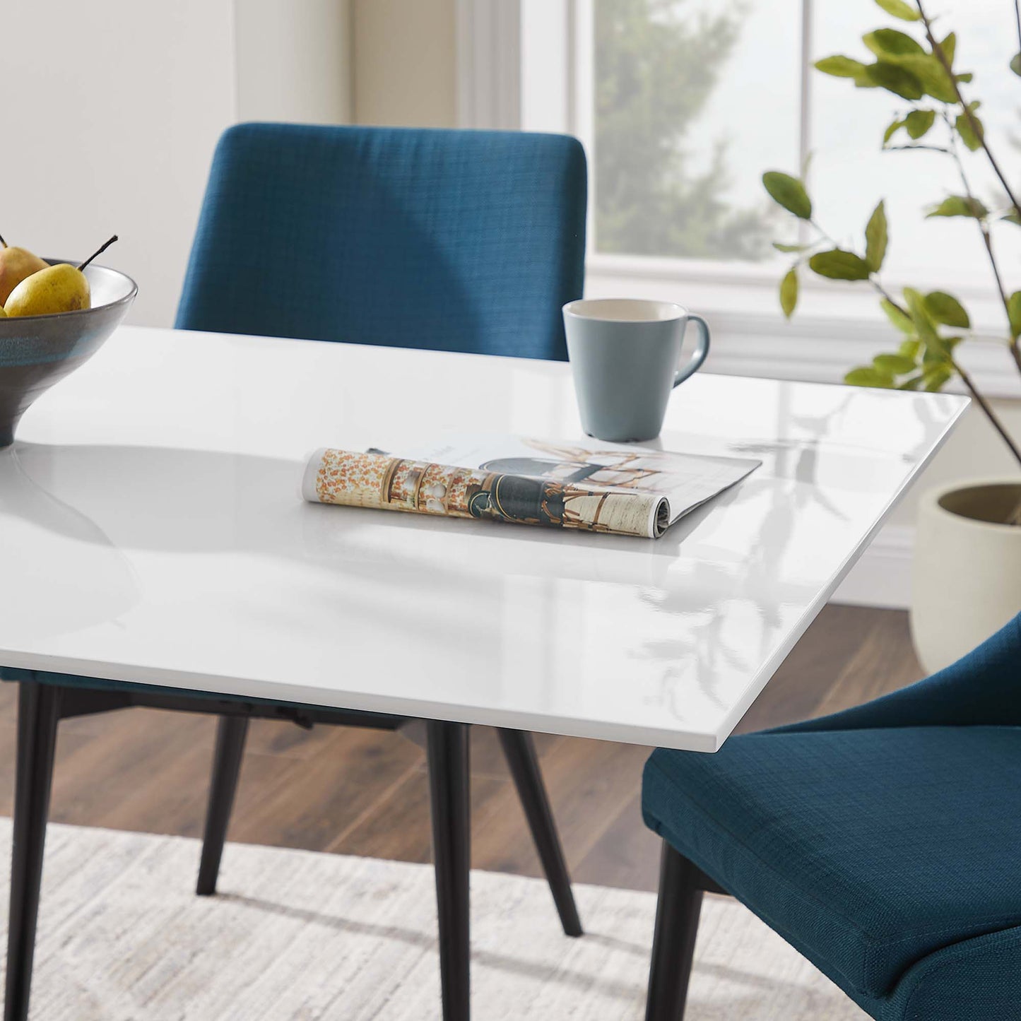 Glossy white dining table with black legs, blue upholstered chairs, mug, rolled newspaper, fruit bowl, and leafy plant.