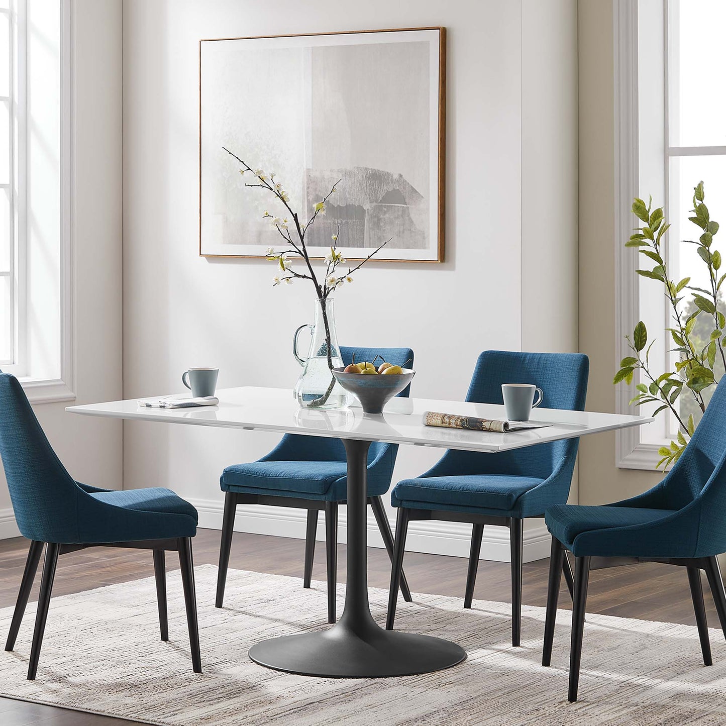 Modern white dining table with pedestal base, blue chairs, vase with branches, fruit bowl, & gray rug.