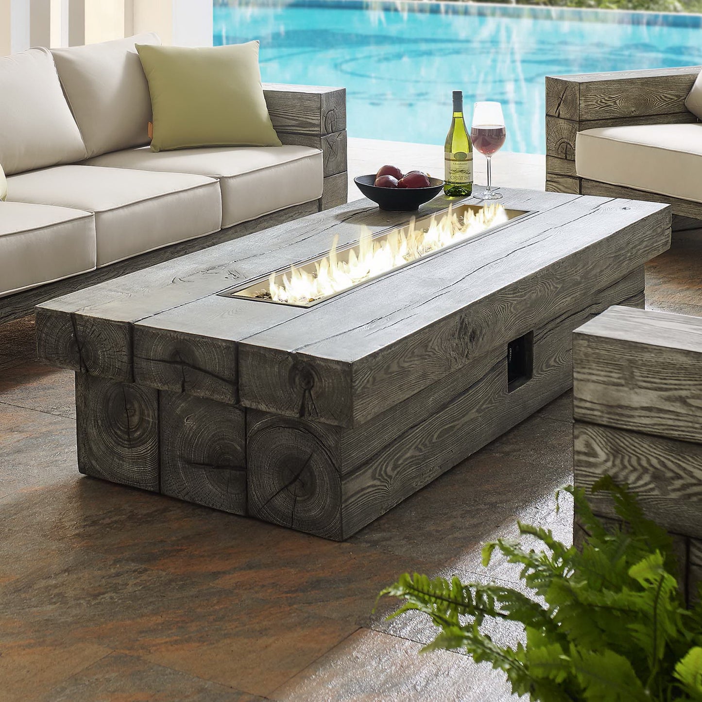Rectangular outdoor fire pit table in light gray with flames, poolside beige sectional sofa, wine bottle, glass and fruit bowl.