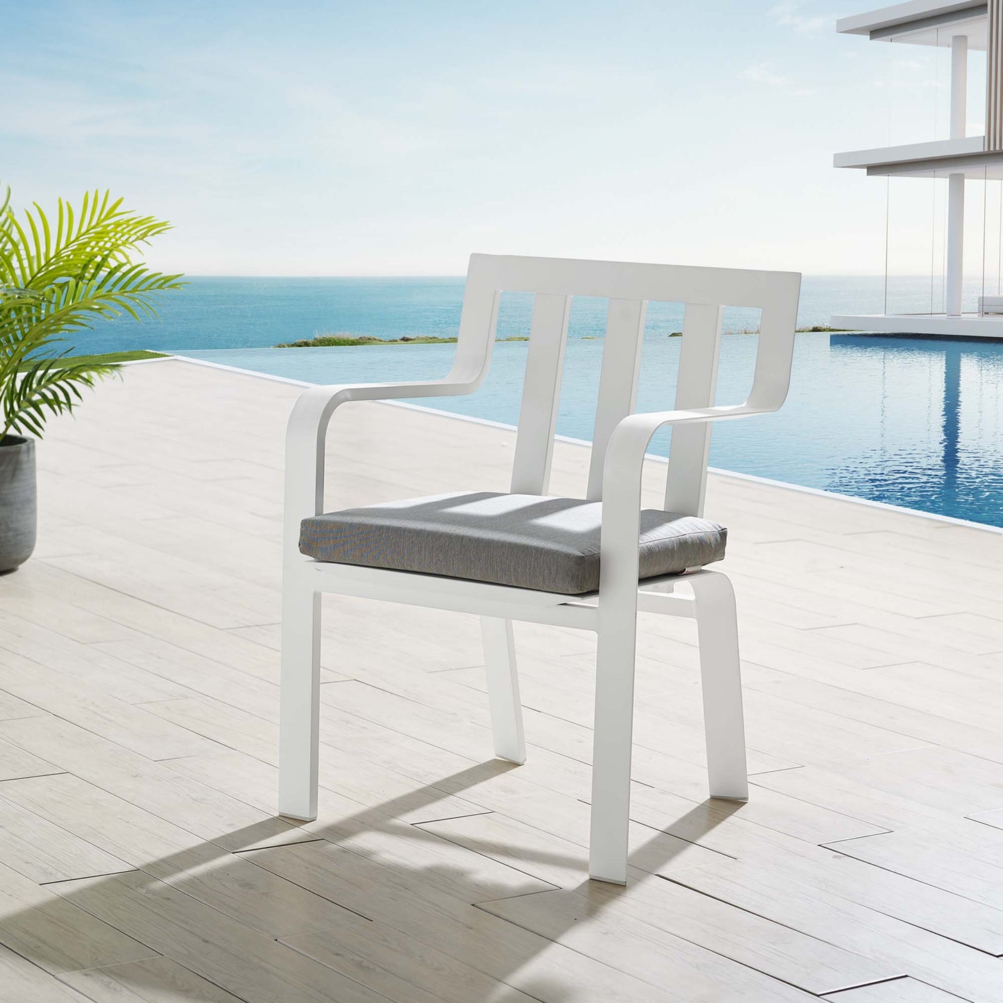 White outdoor patio armchair with curved arms and slatted back, gray cushion, on a wood deck by a pool with an ocean view.