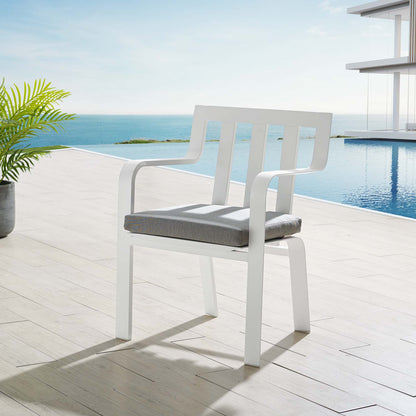 White outdoor patio armchair with curved arms and slatted back, gray cushion, on a wood deck by a pool with an ocean view.